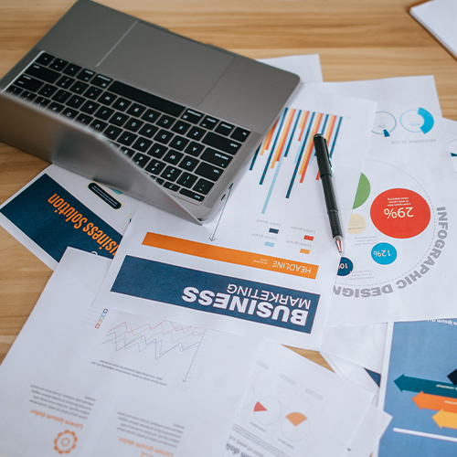 A laptop and scattered business plans, charts and infographics on a wooden desk.